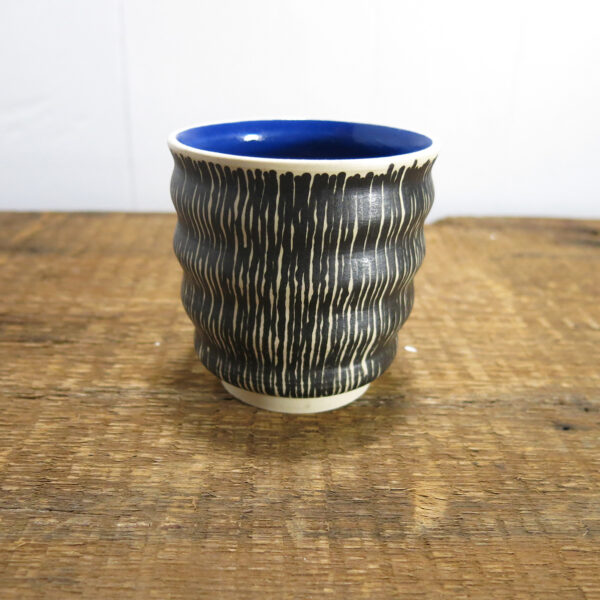 Small ceramic cup made with white clay, with undulating walls, black lines outside, and blue inside.