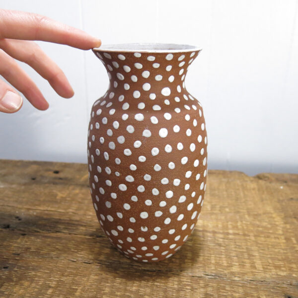 A hand touching a Red clay vase with white spots.