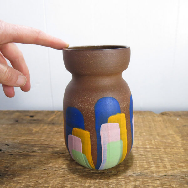A hand touching a red clay vase with colorful stripes.