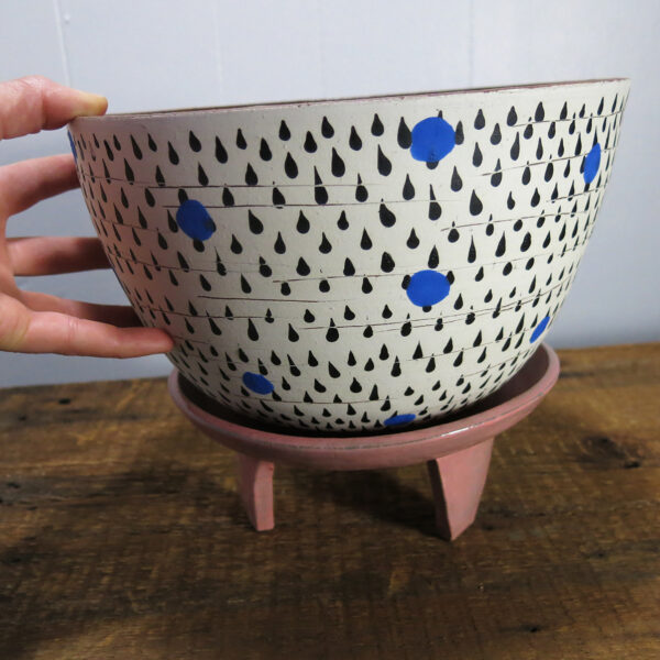 A hand appears to pick up a large ceramic planter with black and blue decoration, sitting on a pink, three-legged ceramic stand.
