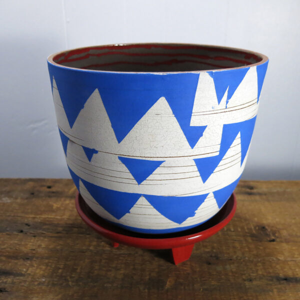 A slightly different view of a blue and white ceramic planter on a red, three-legged ceramic stand. Red stripes are visible on the interior of the planter.