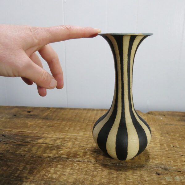 A hand reaching out and touching a black-striped, cream-colored ceramic vase with a very tall neck.