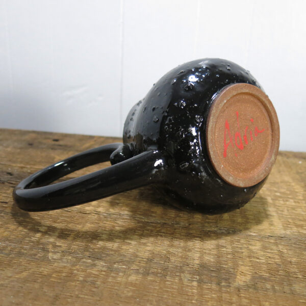 A black ceramic pitcher with a large handle lying on its side, showing the red signature underneath.